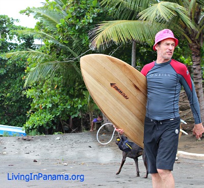 Man with surfboard on beach with trees, boat, dog in background