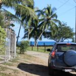 Photo of car on dirt road with palm trees and ocean in distance