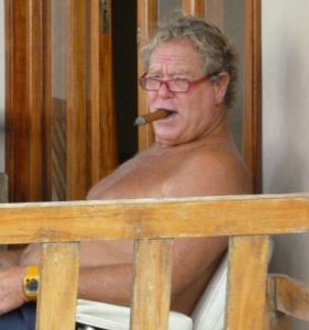 older man looking toward camera. He is sitting bare-chested, smoking a cigar, and wearing glasses