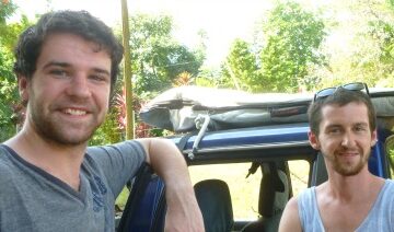 2 men in front of blue pick up with surfboard on top