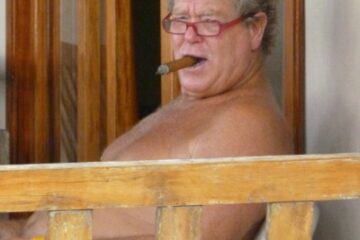 older man looking toward camera. He is sitting bare-chested, smoking a cigar, and wearing glasses