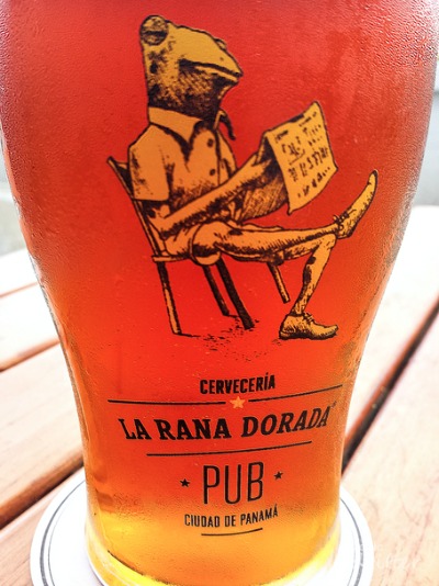 a cold glass of beer with logo of a golden frog sitting in a chair reading the paper