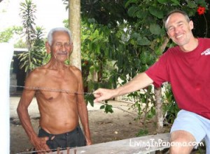 An old man with no shirt next to a middle aged man leaning on a fence