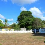 flat grassy lot, white fence, yellow house, trees, blue sky