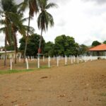 flat dirt area, white posts, trees, 1 house on left and right