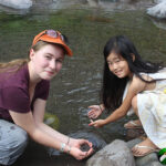 2 girls crouching in river, catching tadpoles